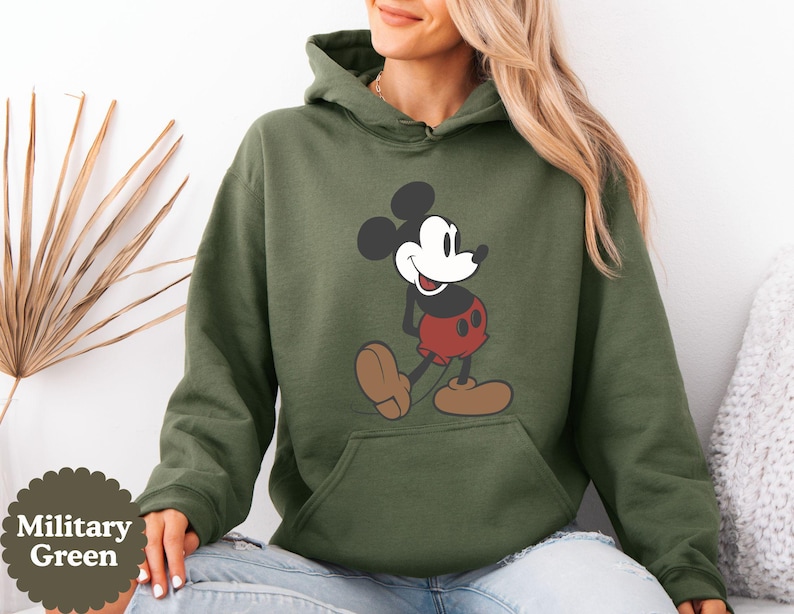 May include: Military green hoodie featuring a classic Mickey Mouse graphic. The cartoon character is depicted in his iconic red shorts, brown shoes, and black and white features. The hoodie has a kangaroo pocket and a drawstring hood.