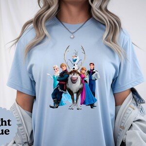 May include: A light blue t-shirt featuring a graphic print of the characters Elsa, Kristoff, Olaf, Sven, Anna, and Hans from the movie Frozen. The shirt has short sleeves and a crew neckline, showcasing the colourful animated figures centred on the chest.