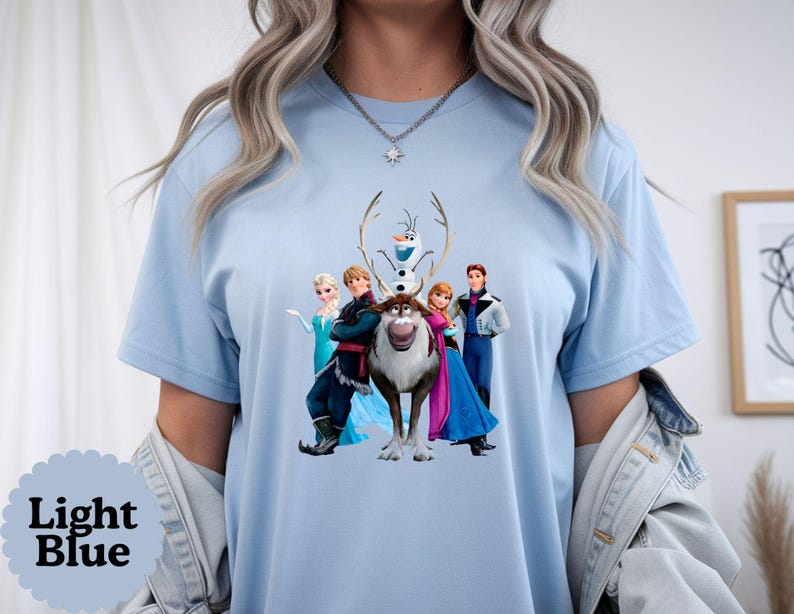 May include: A light blue t-shirt featuring a graphic print of the characters Elsa, Kristoff, Olaf, Sven, Anna, and Hans from the movie Frozen. The shirt has short sleeves and a crew neckline, showcasing the colourful animated figures centred on the chest.