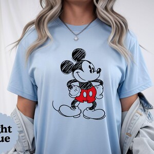 May include: Light blue t-shirt featuring a black and red cartoon character. The character has black ears, a black face, and red shorts. The text "Light Blue" is in a scalloped circle on the bottom left of the shirt.