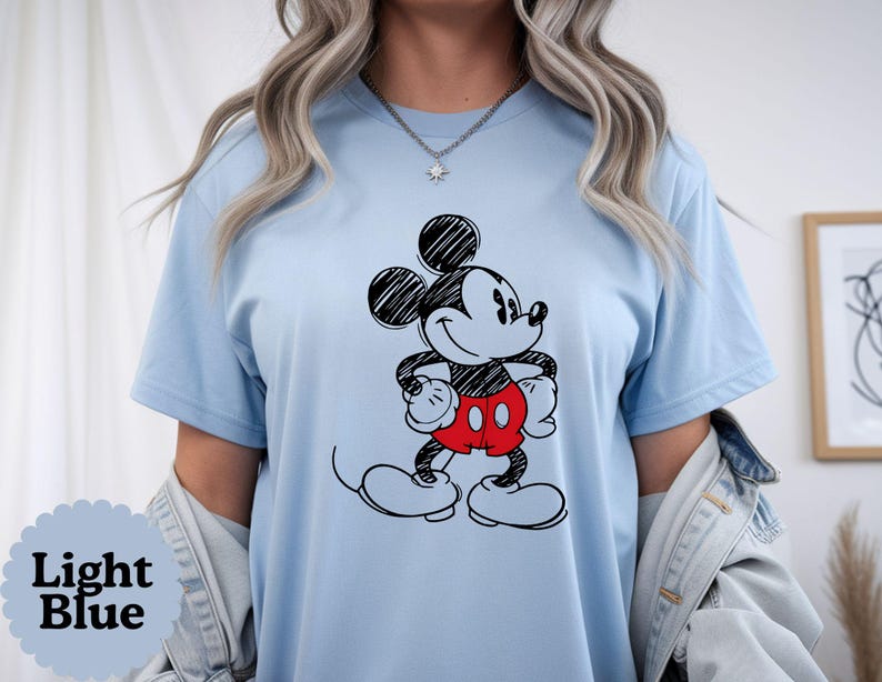 May include: Light blue t-shirt featuring a black and red cartoon character. The character has black ears, a black face, and red shorts. The text "Light Blue" is in a scalloped circle on the bottom left of the shirt.
