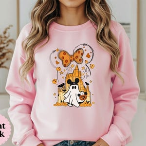 May include: A light pink crewneck sweatshirt featuring a graphic print of a white ghost wearing mouse ears, standing in front of a yellow castle. The design includes an orange polka dot bow, small pumpkins, bats, and stars surrounding the central figure.