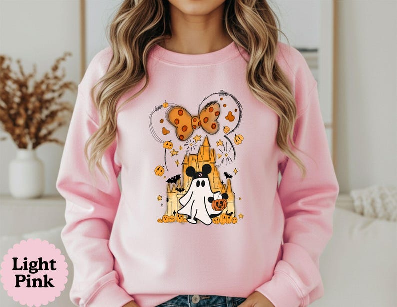 May include: A light pink crewneck sweatshirt featuring a graphic print of a white ghost wearing mouse ears, standing in front of a yellow castle. The design includes an orange polka dot bow, small pumpkins, bats, and stars surrounding the central figure.