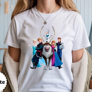 May include: A white short-sleeved t-shirt featuring a graphic print of characters from the movie Frozen, including Elsa, Anna, Kristoff, Sven the reindeer, and Olaf the snowman, arranged in a group centred on the front of the garment.
