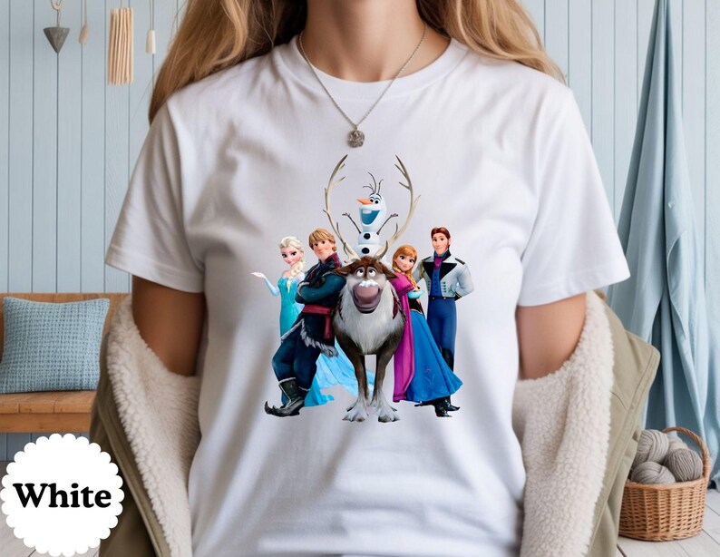 May include: A white short-sleeved t-shirt featuring a graphic print of characters from the movie Frozen, including Elsa, Anna, Kristoff, Sven the reindeer, and Olaf the snowman, arranged in a group centred on the front of the garment.