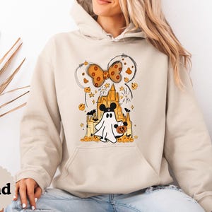 May include: A sand-colored hooded sweatshirt featuring a graphic print of a ghost wearing mouse ears, standing in front of a castle. The design includes a polka-dot bow, small pumpkins, stars, and bats in shades of orange, black, and yellow.
