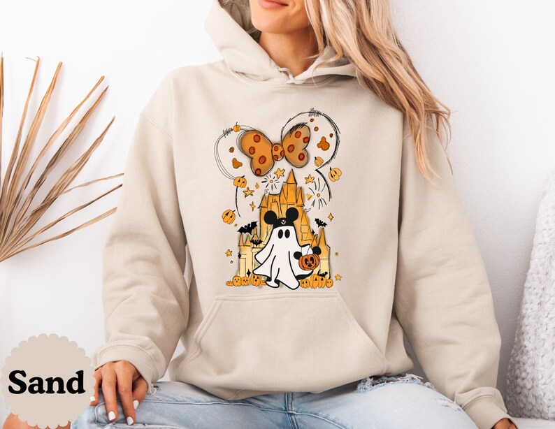 May include: A sand-colored hooded sweatshirt featuring a graphic print of a ghost wearing mouse ears, standing in front of a castle. The design includes a polka-dot bow, small pumpkins, stars, and bats in shades of orange, black, and yellow.