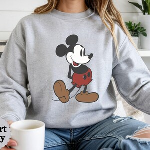 May include: Sport Grey sweatshirt featuring a large graphic of Mickey Mouse in a walking pose. Mickey is depicted in his classic colors: black ears, red shorts, and brown shoes. The sweatshirt is a light gray color.