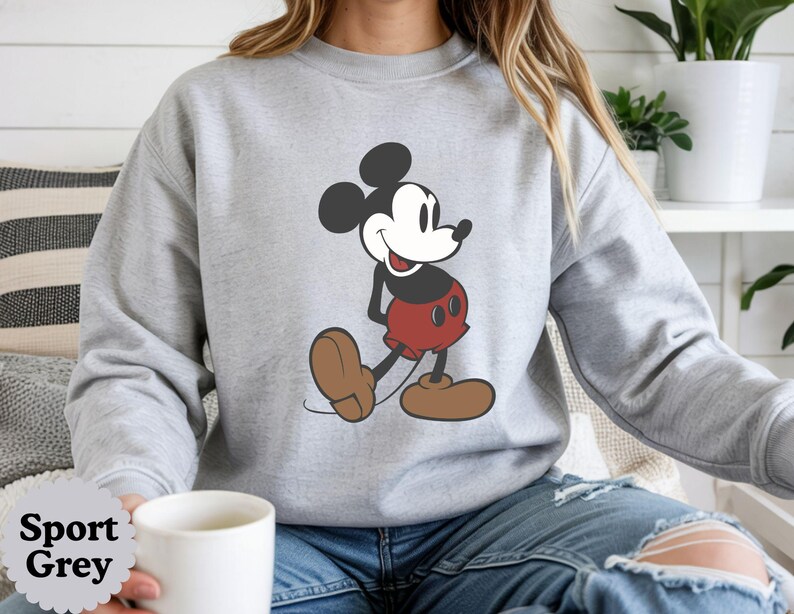 May include: Sport Grey sweatshirt featuring a large graphic of Mickey Mouse in a walking pose. Mickey is depicted in his classic colors: black ears, red shorts, and brown shoes. The sweatshirt is a light gray color.