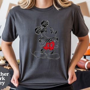 May include: Heather dark grey t-shirt featuring a sketched Mickey Mouse design. The cartoon character is outlined in black with a scribbled effect, wearing red shorts. The shirt is a dark grey color.
