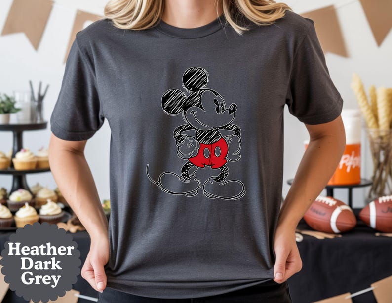 May include: Heather dark grey t-shirt featuring a sketched Mickey Mouse design. The cartoon character is outlined in black with a scribbled effect, wearing red shorts. The shirt is a dark grey color.