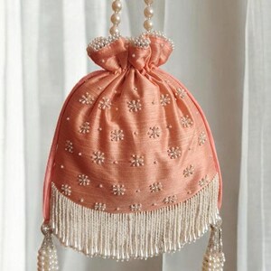 May include: A peach-coloured drawstring bag with a pearl beaded fringe and a pearl chain strap. The bag is decorated with small white flower embroidery.