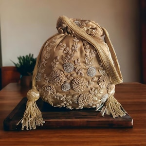 May include: A gold-colored, round drawstring bag with intricate floral embroidery and beaded accents. The bag features a beaded strap and tassel details, resting on a wooden surface. The design includes embroidered flowers and leaves.