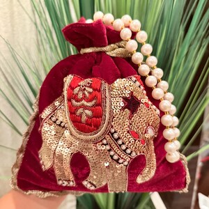 May include: A burgundy velvet pouch with a gold and red embroidered elephant design. The pouch has a pearl string handle.