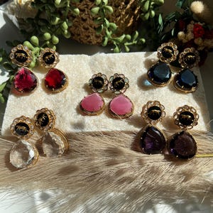 May include: Six pairs of dangle earrings with black and gold accents. Each pair features a different colored gemstone: red, pink, blue, clear, and purple.