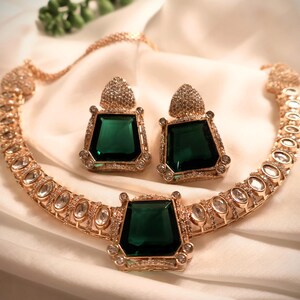 May include: A gold necklace and earrings set with a large emerald green stone in the center of the necklace and smaller emerald green stones in the earrings. The necklace and earrings are adorned with clear rhinestones.