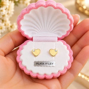 Puede incluir: Un par de pendientes de botón en forma de corazón dorado con una pequeña piedra clara en cada uno, presentados en un joyero rosa con forma de concha, interior blanco y una etiqueta plateada con la inscripción PLATA 1ª LEY.