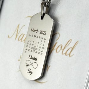 Puede incluir: Collar con placa de identificación de plata con un calendario para marzo de 2025, con el 20 rodeado. Los textos "Charlotte" y "Tony" están grabados debajo del calendario, junto con un símbolo de infinito y un corazón.
