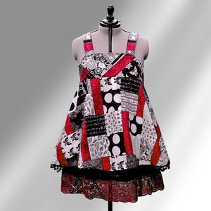 May include: A patchwork dress on a mannequin. The dress combines black, white, and red fabrics with patterns like polka dots and florals. The dress has a tiered skirt with lace trim and black pom-pom trim. The dress is a unique, handmade item.