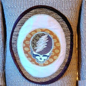 May include: A gray knit sweater dress with a round patch featuring a skull and lightning bolt design. The dress has long sleeves, a ruffled collar, and a ruffled hem. The dress is displayed on a black mannequin.