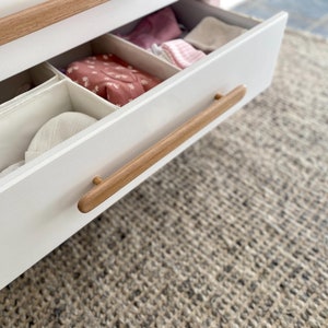 May include: A white dresser drawer with a wooden handle. The drawer is open and contains several fabric storage bins and a white hat.