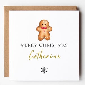 May include: A Christmas card featuring a gingerbread man illustration. The card says "MERRY CHRISTMAS Catherine" in black and gold script, with a snowflake graphic. The card is on a white background with a brown envelope.