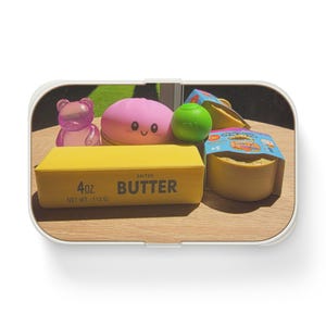 May include: A collection of small toys including a purple translucent bear, a pink round character with a smiling face, a green ball, and a yellow rectangular block labeled 4oz. SALTED BUTTER NET WT. 113 G, alongside a yellow circular container.
