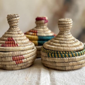 Puede incluir: Tres recipientes tejidos con tapa de color beige natural. Las tapas y las bases están decoradas con detalles rojos, amarillos, verdes y azules. Los recipientes están hechos de un material de fibra natural.