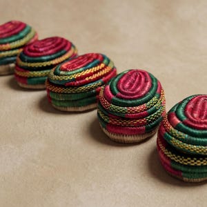 Puede incluir: Cinco pequeñas cajas decorativas de forma ovalada. Cada caja está bordada intrincadamente con hilos coloridos en tonos rojos, verdes, amarillos y naranjas. Las cajas están dispuestas en diagonal sobre una superficie de tono neutro.