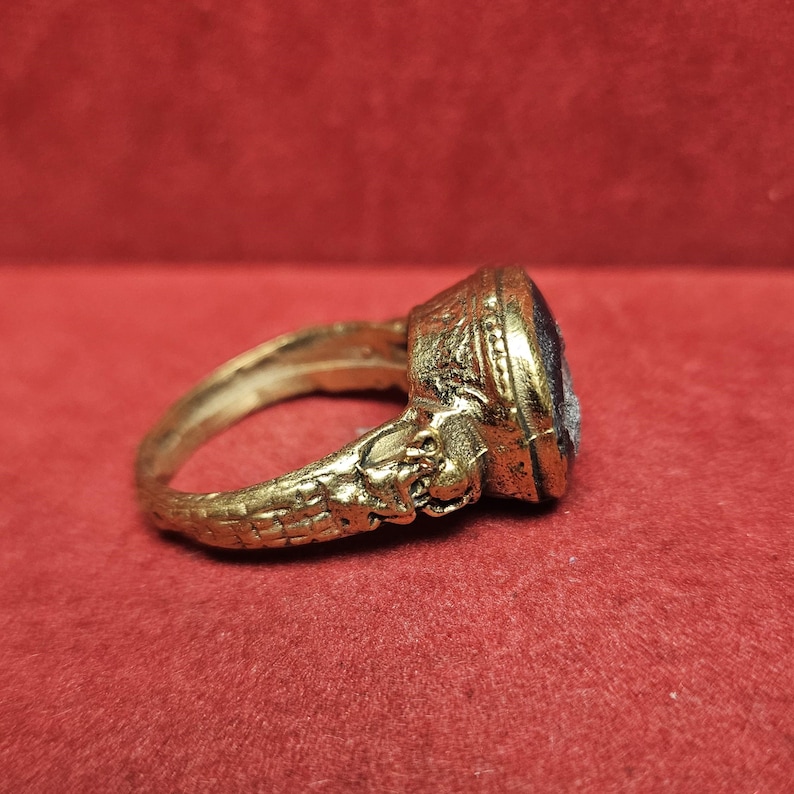 May include: A gold-colored ring with an oval, dark-colored stone. The ring has intricate detailing, including a textured band and sculpted elements near the stone. The ring is set against a red background.