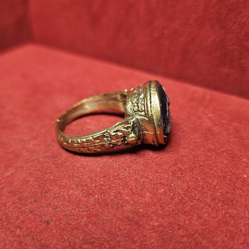 May include: A gold-toned ring with an oval, dark-colored stone. The ring features intricate detailing, including a textured band and ornate setting around the stone. The ring is displayed against a red background.