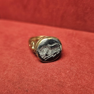 May include: A gold ring with an oval, black stone featuring a white engraved image of a running animal. The ring has ornate detailing on the band and around the stone setting. The ring is set against a red background.