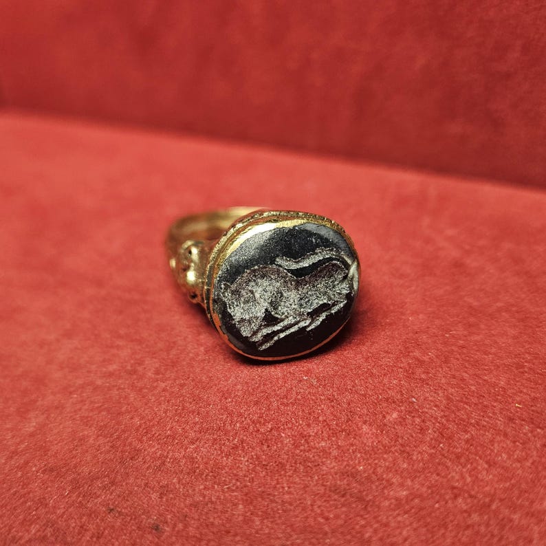 May include: A gold ring with an oval, black stone featuring a white engraved image of a running animal. The ring has ornate detailing on the band and around the stone setting. The ring is set against a red background.