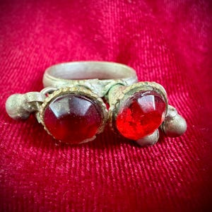 Może przedstawiać: Antyczny pierścionek w kolorze srebrnym z dwoma wisiorkami, każdy z dużym, fasetowanym, czerwonym kamieniem. Pierścionek i wisiorki spoczywają na głębokiej czerwonej aksamitnej powierzchni, tworząc uderzający kontrast.