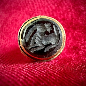 May include: A circular gold ring featuring a black stone inlay. The centre of the stone displays a detailed, light-coloured intaglio carving of a four-legged animal in profile, facing right, with its head turned back towards its body.