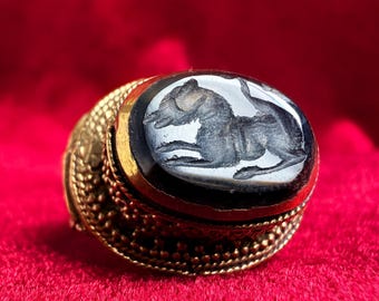 May include: A gold ring featuring an oval-shaped black stone inlay with a carved relief of a four-legged animal. The gold band displays a textured, beaded surface pattern surrounding the central stone setting.