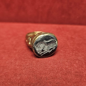 May include: A gold-colored ring with a dark, oval-shaped stone. The stone features a carved image of a running animal. The ring is set against a red background, highlighting the ring's details and design.