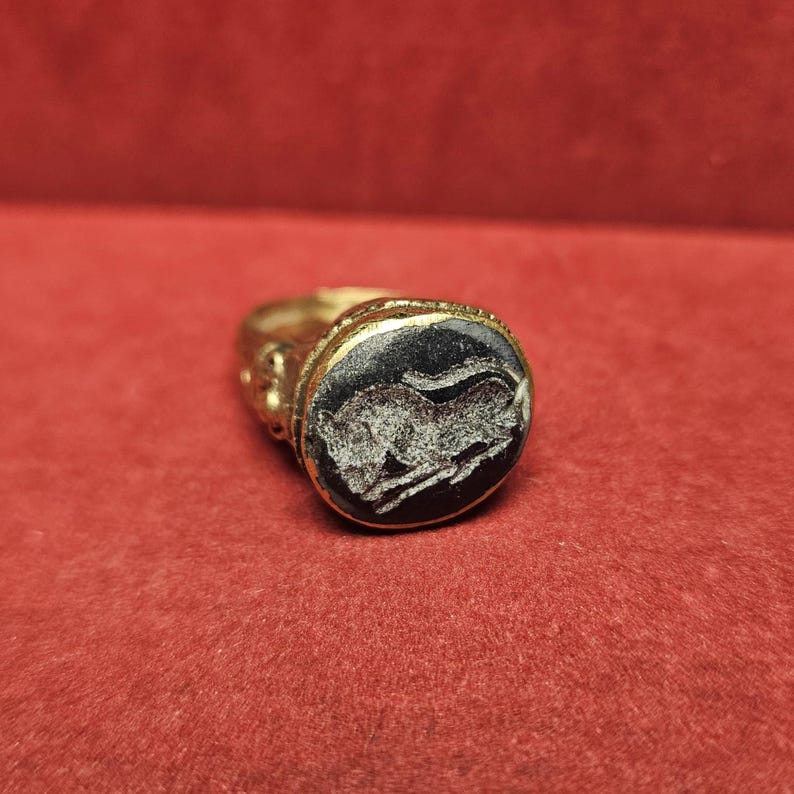 May include: A gold-colored ring with a dark, oval-shaped stone. The stone features a carved image of a running animal. The ring is set against a red background, highlighting the ring's details and design.