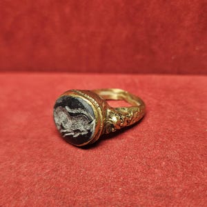 May include: A gold ring with a dark, possibly onyx, stone. The stone features a white, possibly carved, design. The ring's band has detailed patterns. The ring is set against a red background.