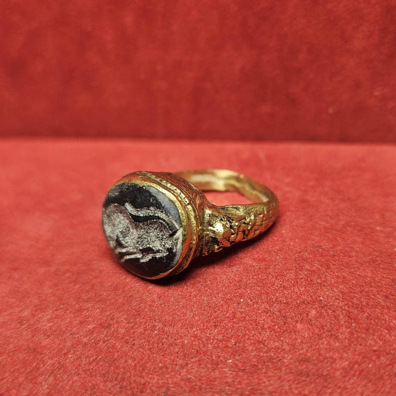 May include: A gold ring with a dark, possibly onyx, stone. The stone features a white, possibly carved, design. The ring's band has detailed patterns. The ring is set against a red background.