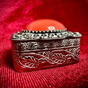 Könnte beinhalten: Eine rechteckige Metallschatulle mit polierter silberfarbener Oberfläche und geprägten Blattmustern an den Seiten. Der Deckel ist mit einem glatten, abgerundeten, leuchtend orangefarbenen Cabochon-Stein verziert, der in einem gewellten Rand sitzt.