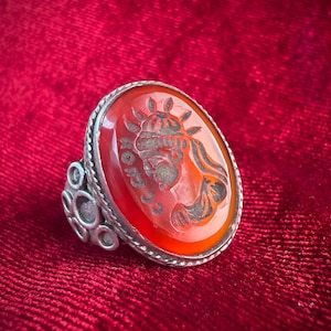 May include: A silver ring featuring a large, oval-shaped, translucent orange carnelian gemstone. The stone is engraved with a profile of a figure wearing a crown and includes text characters. The ring band has a decorative, multi-circle design.