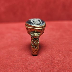 May include: A gold-colored ring with an oval, dark blue stone. The stone features a white, detailed carving. The ring's band and setting are intricately designed with ornate patterns and figures. The ring is set against a red background.