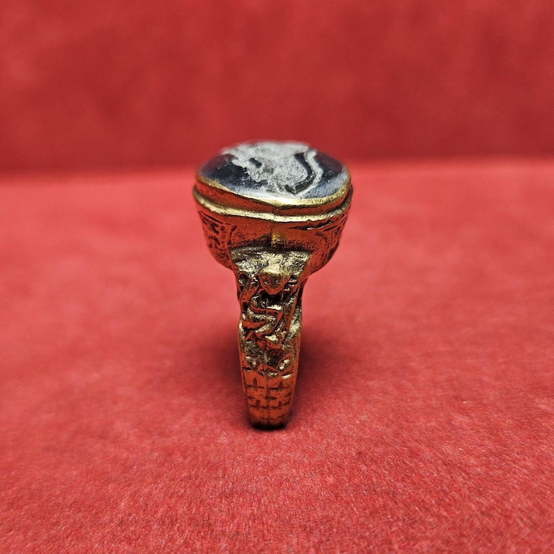May include: A gold-colored ring with an oval, dark blue stone. The stone features a white, detailed carving. The ring's band and setting are intricately designed with ornate patterns and figures. The ring is set against a red background.
