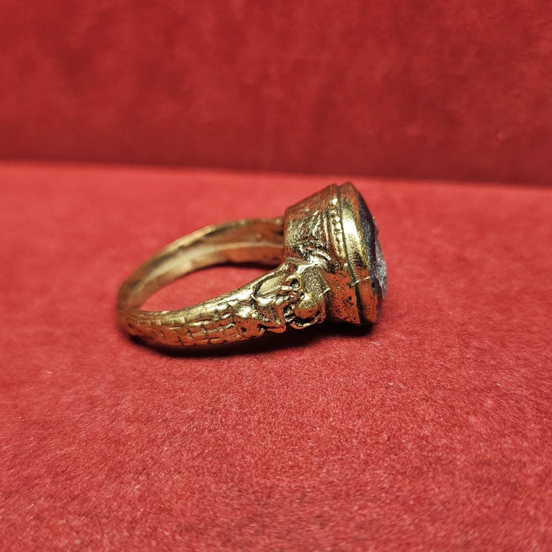 May include: A gold-colored ring with an oval, dark blue stone. The ring has intricate detailing on the band and around the stone setting. The ring is displayed on a red surface.