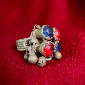Könnte beinhalten: Ein metallischer Ring mit einem strukturierten Band und einer Ansammlung hängender Elemente. Das Design umfasst mehrere konische Fassungen mit abgerundeten blauen und roten Steinen, durchsetzt mit kleinen, kugelförmigen Metallanhängern an Ösen.