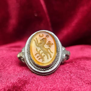 May include: Antique silver ring with an oval, amber-colored stone. The stone features a carved design of a deer, crescent moon, and star. The ring has a detailed, rope-like border and decorative shoulders.