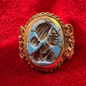 Op de afbeelding: Een goudkleurige metalen ring met een ovaalvormige, lichtblauwe steen met een donker, ingesneden profielportret. De ringband heeft een ingewikkeld, getextureerd en gevlochten metaalontwerp rondom de centrale zetting van de steen.