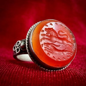 Könnte beinhalten: Ein Silberring mit einem runden, orangefarbenen Karneol-Edelstein als Intaglio. Der Stein ist mit einer Darstellung eines springenden Löwen und eines kleineren Tieres darunter graviert. Der Metallring hat einen gedrehten Rand.