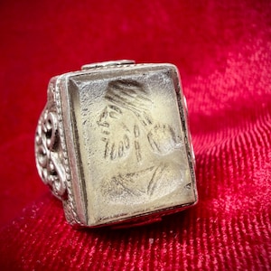 Könnte beinhalten: Silberring mit rechteckiger Oberfläche, die ein geschnitztes Profil einer Person mit Kopfschmuck zeigt. Der Ring hat verzierte Details an den Seiten und ist vor rotem Hintergrund platziert.